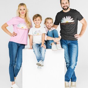 May include: A family of four wearing matching t-shirts with a daisy and the text "DaisyApparelArt". The family is standing in front of a white background. The father is wearing a dark grey t-shirt, the mother is wearing a pink t-shirt, and the two children are wearing white t-shirts.