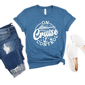 May include: A blue t-shirt with a white graphic that says "On Cruise Control" with a cruise ship, anchor, and star. The shirt is paired with blue jeans and white striped slip-on shoes.