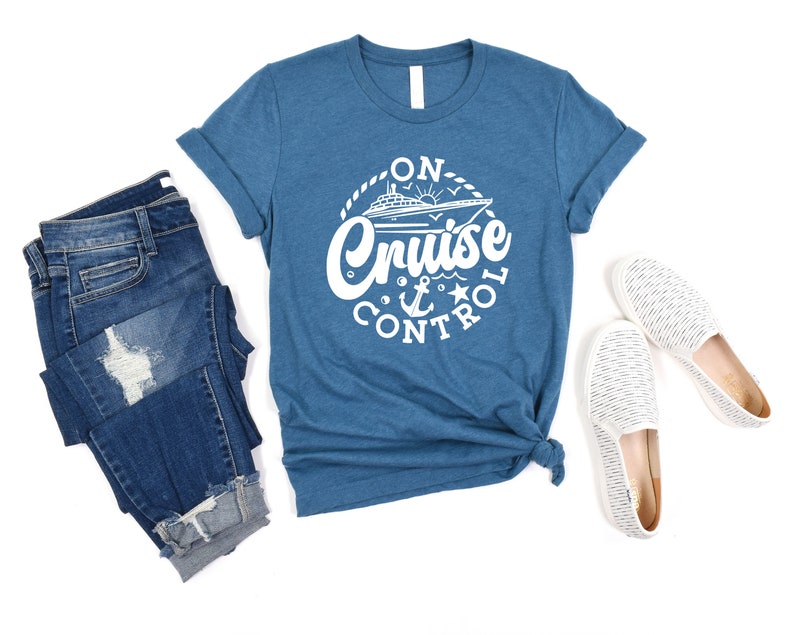 May include: A blue t-shirt with a white graphic that says "On Cruise Control" with a cruise ship, anchor, and star. The shirt is paired with blue jeans and white striped slip-on shoes.