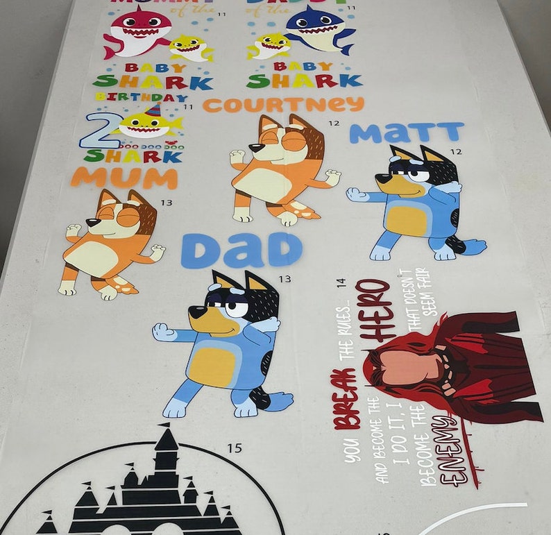 May include: A collection of colorful cartoon character decals, including sharks, dogs, and a superhero. Text includes "Baby Shark," "Birthday," and names. The decals are designed for various applications.