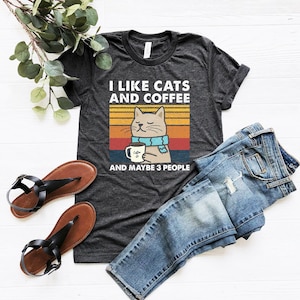 May include: A dark grey t-shirt with a retro-style graphic of a cat drinking coffee. The text on the shirt reads "I like cats and coffee and maybe 3 people".