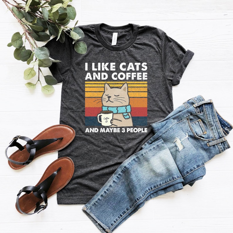 May include: A dark grey t-shirt with a retro-style graphic of a cat drinking coffee. The text on the shirt reads "I like cats and coffee and maybe 3 people".