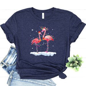 May include: A navy blue t-shirt with a Christmas-themed design featuring two pink flamingos wearing Santa hats and adorned with colorful Christmas lights. The flamingos are standing in a snowy scene.