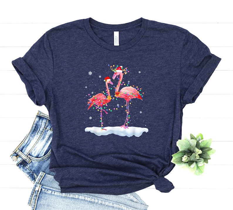 May include: A navy blue t-shirt with a Christmas-themed design featuring two pink flamingos wearing Santa hats and adorned with colorful Christmas lights. The flamingos are standing in a snowy scene.