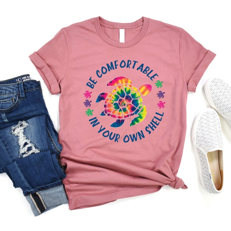 Może przedstawiać: R&oacute;żowa koszulka z podwiniętymi rękawami, z wzorem ż&oacute;łwia tie-dye i napisem &bdquo;BE COMFORTABLE IN YOUR OWN SHELL&rdquo;. Koszulka jest połączona z niebieskimi dżinsami i białymi butami wsuwanymi.