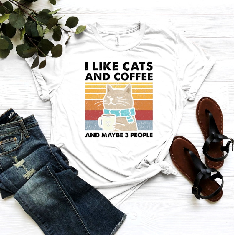 May include: White t-shirt featuring a retro-style graphic of a cat holding a cup of coffee. The text reads "I like cats and coffee and maybe 3 people".