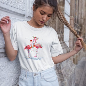 May include: White t-shirt with a Christmas-themed design featuring two pink flamingos with colorful lights wrapped around them. The flamingos are wearing Santa hats.