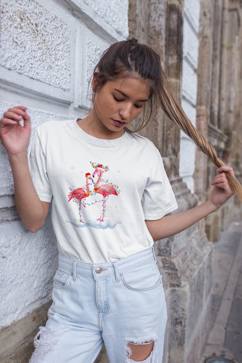 May include: White t-shirt with a Christmas-themed design featuring two pink flamingos with colorful lights wrapped around them. The flamingos are wearing Santa hats.