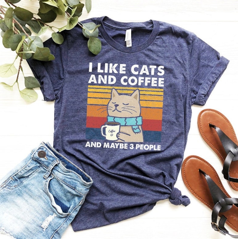 May include: A navy blue t-shirt featuring a retro-style graphic of a cat wearing a scarf and holding a cup of coffee. The text on the shirt reads "I like cats and coffee and maybe 3 people".