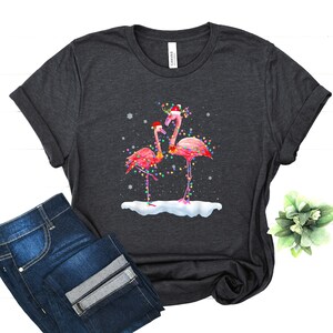 May include: A dark grey t-shirt with a Christmas-themed design featuring two pink flamingos wearing Santa hats and surrounded by Christmas lights. The flamingos are standing on a snowy surface.