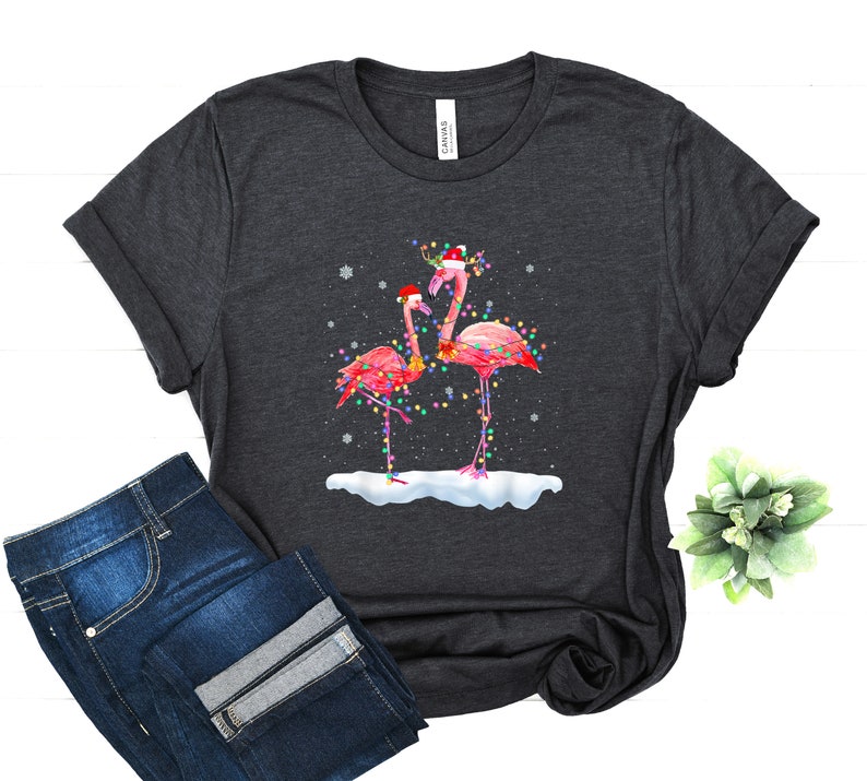 May include: A dark grey t-shirt with a Christmas-themed design featuring two pink flamingos wearing Santa hats and surrounded by Christmas lights. The flamingos are standing on a snowy surface.