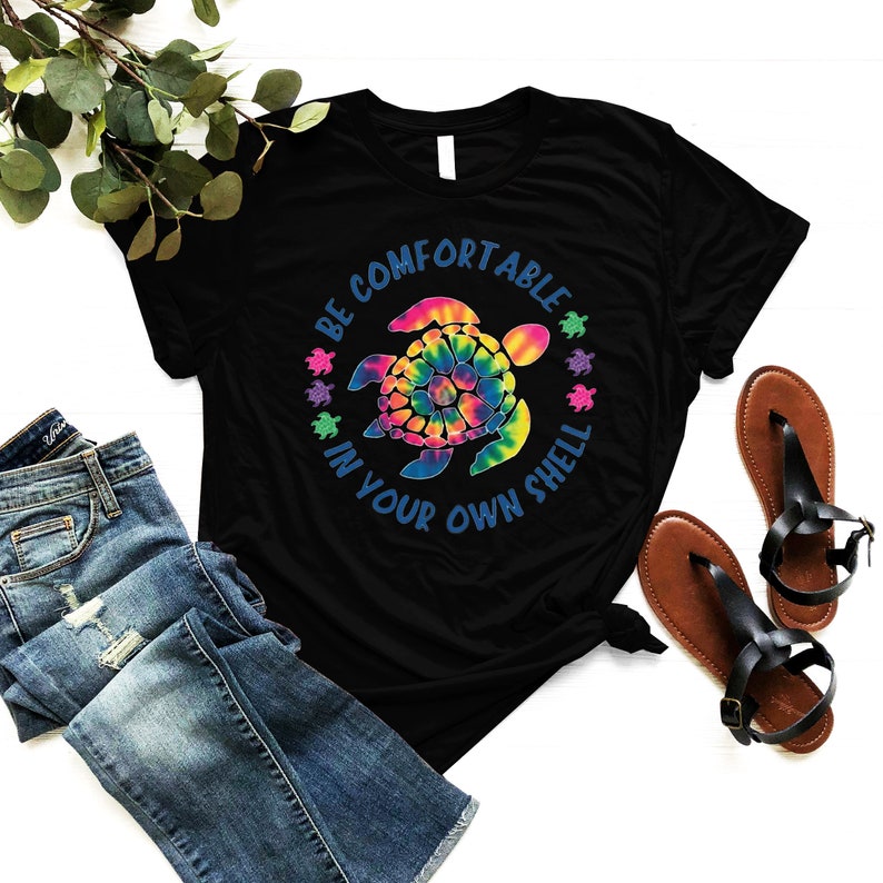 Może przedstawiać: Czarny t-shirt z wzorem ż&oacute;łwia tie-dye i napisem "BE COMFORTABLE IN YOUR OWN SHELL" w kolorze niebieskim. Ż&oacute;łw jest wielokolorowy. Koszulka jest połączona z niebieskimi dżinsami i brązowymi sandałami.