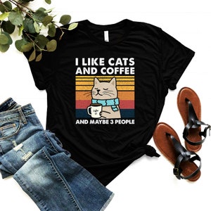 May include: Black t-shirt featuring a retro-style graphic of a cat holding a cup of coffee. The text reads "I like cats and coffee and maybe 3 people".