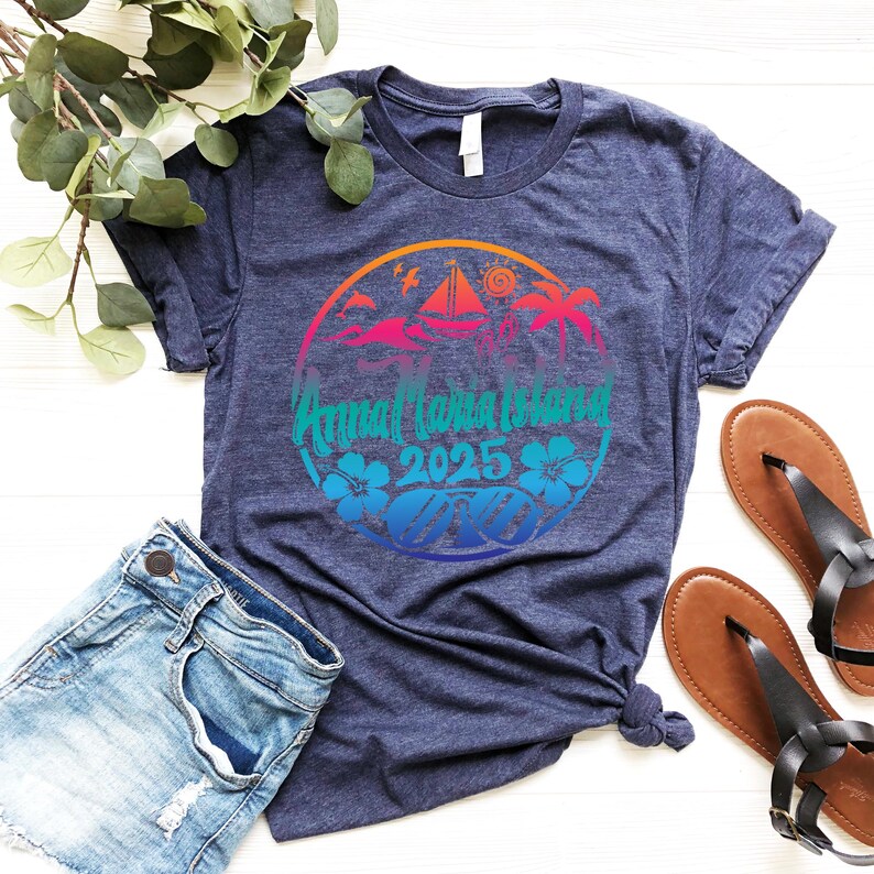 Puede incluir: Una camiseta azul marino con un gr&aacute;fico circular de colores que dice "Annual Banana Island 2025". El gr&aacute;fico presenta palmeras, un velero y una puesta de sol.