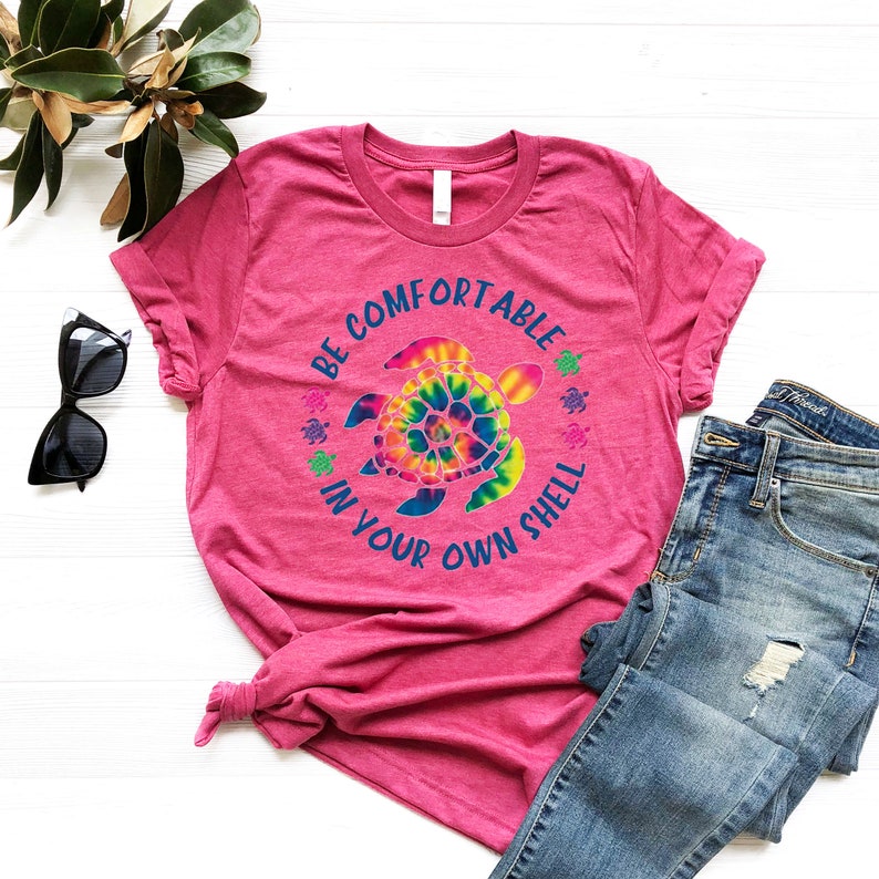 Może przedstawiać: R&oacute;żowa koszulka z napisem "BE COMFORTABLE IN YOUR OWN SHELL" otaczającym wz&oacute;r ż&oacute;łwia tie-dye. Obraz zawiera niebieskie dżinsy i okulary przeciwsłoneczne.