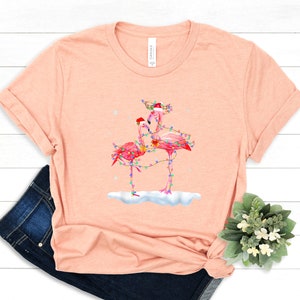 May include: A pink t-shirt with a Christmas-themed design featuring two pink flamingos wearing Santa hats and wrapped in Christmas lights. The flamingos are standing on a snowy surface.