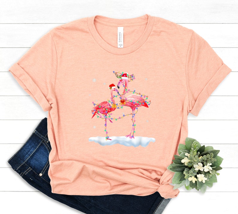 May include: A pink t-shirt with a Christmas-themed design featuring two pink flamingos wearing Santa hats and wrapped in Christmas lights. The flamingos are standing on a snowy surface.