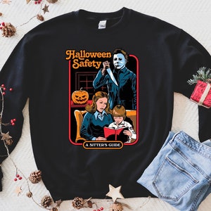 May include: Black sweatshirt featuring a graphic print of a horror movie scene. The graphic depicts a woman and child reading a book titled "Halloween Safety: A Sitter's Guide" with a masked figure standing behind them holding a knife.