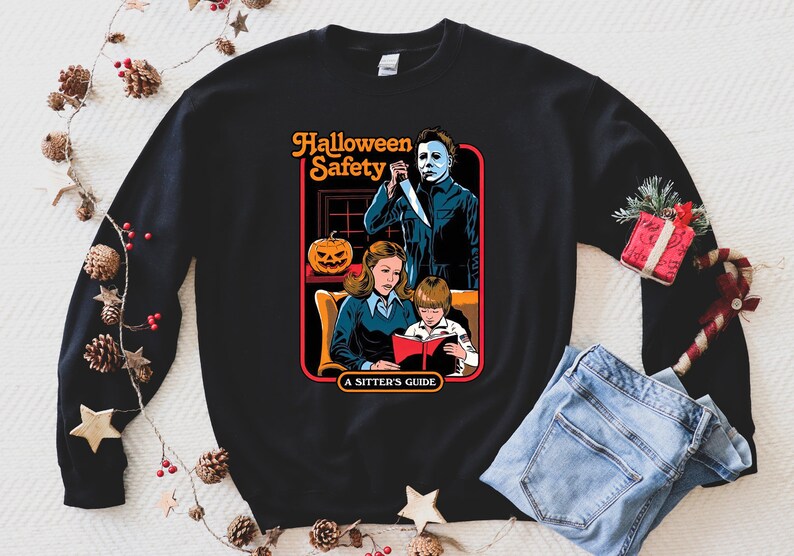 May include: Black sweatshirt featuring a graphic print of a horror movie scene. The graphic depicts a woman and child reading a book titled "Halloween Safety: A Sitter's Guide" with a masked figure standing behind them holding a knife.