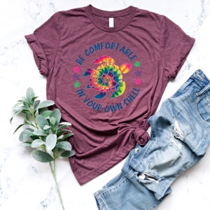 Może przedstawiać: Bordowa koszulka z nadrukiem ż&oacute;łwia morskiego w stylu tie-dye i napisem "BE COMFORTABLE IN YOUR OWN SHELL". Koszulka jest połączona z niebieskimi dżinsami. Gałązka zielonych liści znajduje się w lewym dolnym rogu.