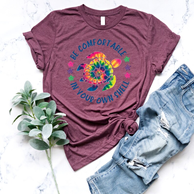 Może przedstawiać: Bordowa koszulka z nadrukiem ż&oacute;łwia morskiego w stylu tie-dye i napisem "BE COMFORTABLE IN YOUR OWN SHELL". Koszulka jest połączona z niebieskimi dżinsami. Gałązka zielonych liści znajduje się w lewym dolnym rogu.