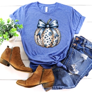 K&ouml;nnte beinhalten: Ein blaues T-Shirt mit einem K&uuml;rbisdesign, einer blauen Schleife und einem floralen Muster. Das Bild zeigt auch braune Stiefeletten und Jeansshorts, wodurch ein herbstliches Ensemble entsteht.