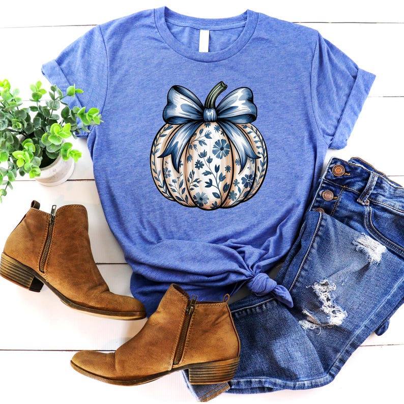 K&ouml;nnte beinhalten: Ein blaues T-Shirt mit einem K&uuml;rbisdesign, einer blauen Schleife und einem floralen Muster. Das Bild zeigt auch braune Stiefeletten und Jeansshorts, wodurch ein herbstliches Ensemble entsteht.