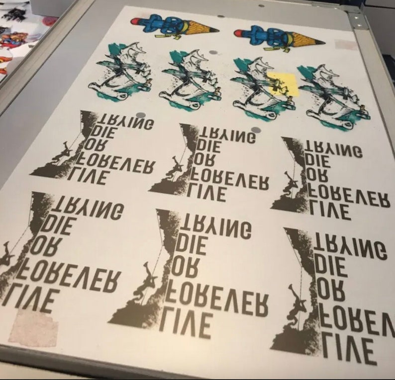 May include: A screen printing of a design with multiple repeats. The design includes a climber, a mountain, and the text "Climb Die OK Love Life". Above are cartoon images of a pencil with a rocket.