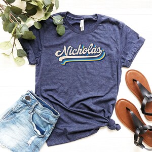 Pode incluir: Uma camiseta azul marinho com um gr&aacute;fico de estilo retr&ocirc; que diz "Nicholas" em cor creme com acentos azuis e amarelos.