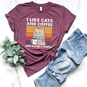 May include: A maroon t-shirt featuring a retro-style graphic of a cat holding a cup of coffee. The text on the shirt reads "I like cats and coffee and maybe 3 people".