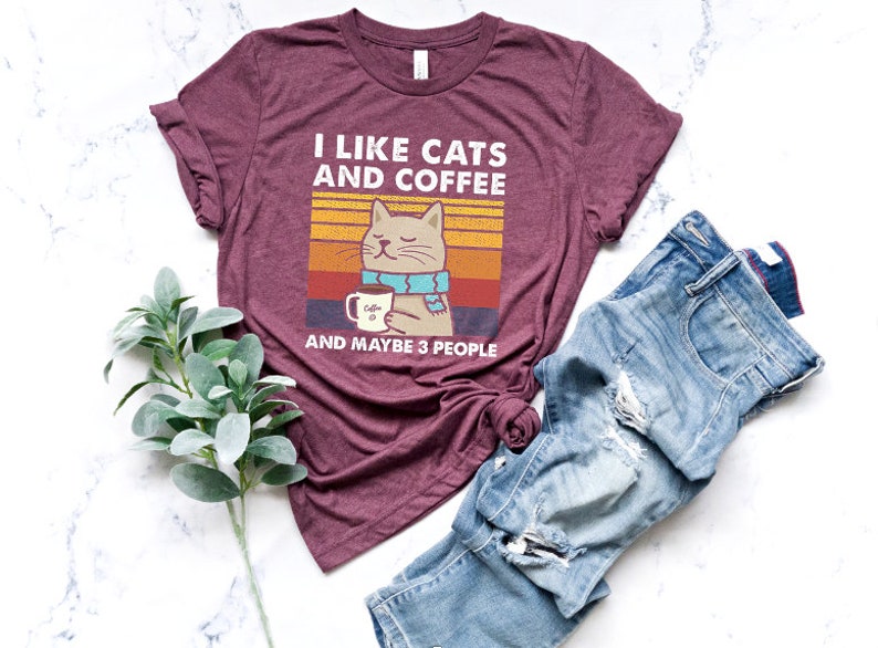 May include: A maroon t-shirt featuring a retro-style graphic of a cat holding a cup of coffee. The text on the shirt reads "I like cats and coffee and maybe 3 people".