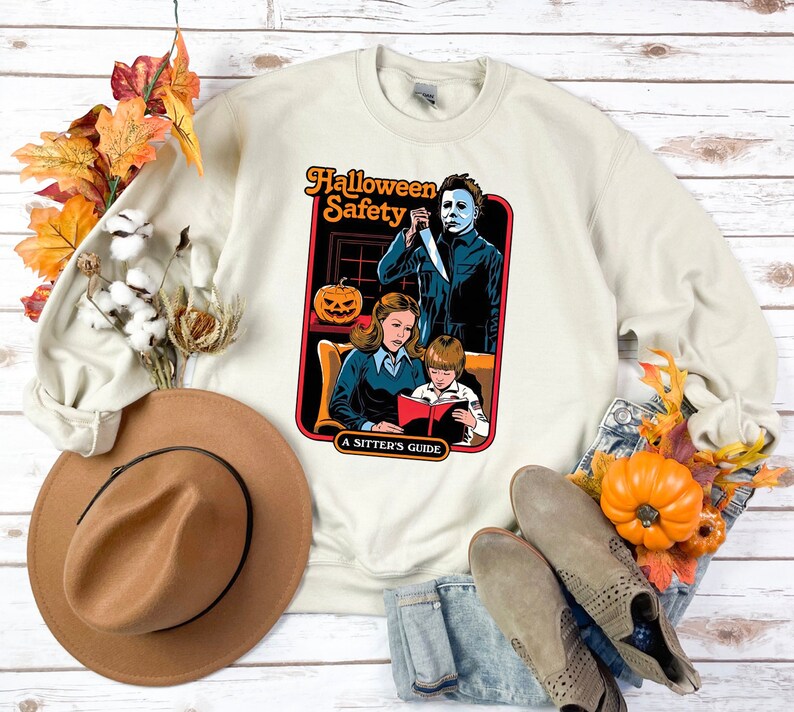 May include: A cream coloured sweatshirt with a graphic of a horror movie scene. The graphic is a book cover with the title "Halloween Safety: A Sitters Guide". The cover features a woman reading a book to a child while a man with a knife stands behind them.