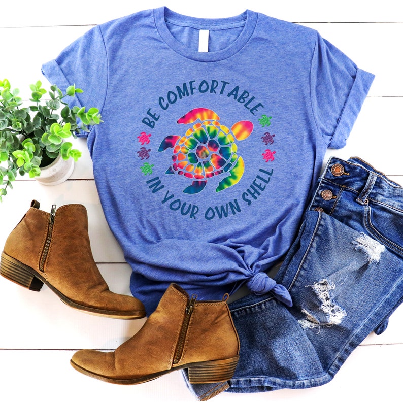 Może przedstawiać: Niebieska koszulka z wzorem ż&oacute;łwia tie-dye i napisem "BE COMFORTABLE IN YOUR OWN SHELL". Na zdjęciu znajdują się dżinsy i brązowe botki z zamkami błyskawicznymi. Widoczna jest r&oacute;wnież mała roślina doniczkowa.