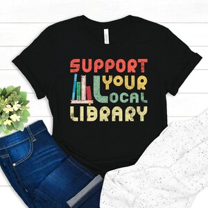 May include: Black t-shirt with a vintage-style graphic that says "Support Your Local Library" with a stack of books in the middle of the text.