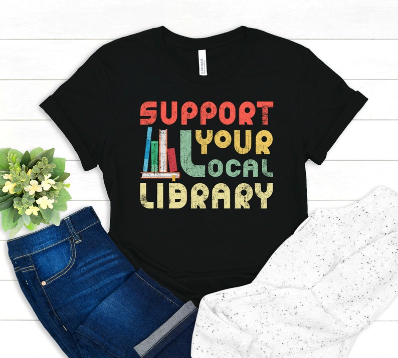 May include: Black t-shirt with a vintage-style graphic that says "Support Your Local Library" with a stack of books in the middle of the text.