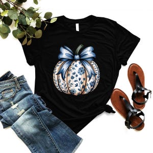 K&ouml;nnte beinhalten: Schwarzes T-Shirt mit einem K&uuml;rbisdesign mit blauer Schleife und floralem Muster. Der K&uuml;rbis hat einen cremefarbenen Hintergrund mit blauen floralen Akzenten. Das Shirt wird mit blauen Jeans und braunen Sandalen kombiniert.