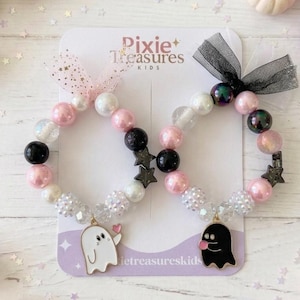 May include: Two beaded bracelets on a Pixie Treasures Kids display card. The left bracelet features pink, white, and black beads with a white ghost charm holding a pink heart. The right bracelet has similar beads with a black ghost charm and a black tulle bow.