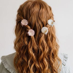 Peut inclure: Cheveux roux longs et ondulés coiffés avec plusieurs pinces à cheveux en forme de coquillage. Les pinces sont dans des tons de rose et de blanc, ajoutant une touche côtière à la coiffure. La personne porte une chemise vert clair.