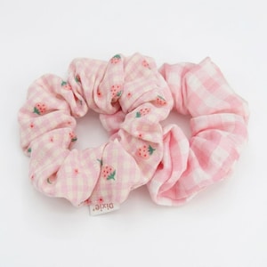Peut inclure: Deux chouchous roses. L'un présente un motif vichy blanc et rose avec des illustrations de fraises. L'autre a un motif vichy rose uni. Accessoires pour cheveux.