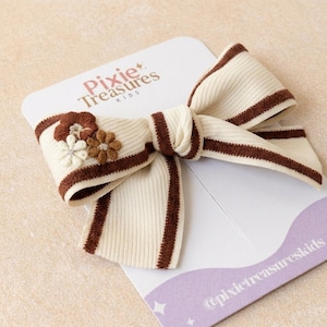 May include: A cream-colored ribbed fabric hair bow featuring dark brown striped edges and three small embroidered flowers in brown and white. The bow is attached to a white display card printed with the text Pixie Treasures KIDS.
