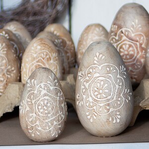 Könnte beinhalten: Eine Sammlung von Holzeiern mit aufwendigen weißen Blumenmustern, die in die Oberfläche geschnitzt sind. Die Eier sind in einem Eierkarton aus Pappe angeordnet.