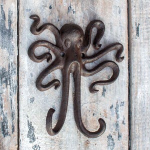 Könnte beinhalten: Ein Wandhaken aus Gusseisen in Form eines Oktopus mit einem rustikalen, verwitterten Finish. Der Oktopus hat acht sich kräuselnde Tentakel und einen detaillierten Kopf. Der Haken ist auf einer verwitterten, hellen Holzoberfläche montiert.