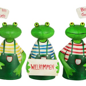 Könnte beinhalten: Drei grüne Froschfiguren in grünen Latzhosen über gestreiften Hemden. Die linken und rechten Frösche halten Schilder mit der Aufschrift Bin im Garten. Der mittlere Frosch hält ein Schild mit der Aufschrift WILLKOMMEN. Große gelbe Augen.