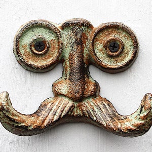 Könnte beinhalten: Ein Wandhaken aus Gusseisen mit einem skurrilen Design. Der Haken hat ein Gesicht mit großen Augen, einen Schnurrbart und zwei Haken zum Aufhängen von Gegenständen. Der Haken ist in Braun- und Grüntönen lackiert.
