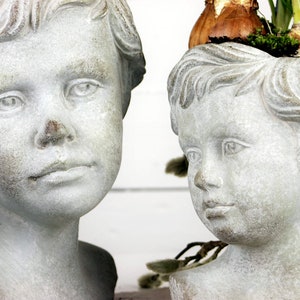 Könnte beinhalten: Zwei verwitterte, graue Steinbüsten von Kindern. Eine Büste hat eine Zwiebel und Grünzeug anstelle eines Kopfes. Die Büsten haben eine abgenutzte Oberfläche, die auf ein antikes oder Gartendekor-Thema hindeutet.