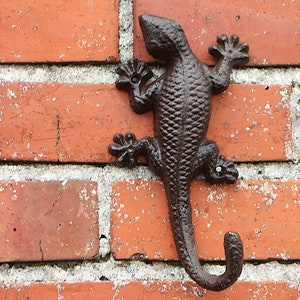 Könnte beinhalten: Ein schwarzer Wandhaken aus Gusseisen in Form einer Eidechse mit einem eingerollten Schwanz. Der Haken ist an einer roten Backsteinwand befestigt.