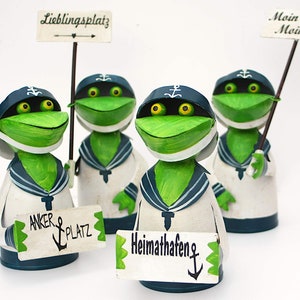 Könnte beinhalten: Vier grüne Froschfiguren, die Matrosenanzüge tragen und Schilder halten. Die Schilder zeigen "Lieblingsplatz", "Anker Platz", "Heimathafen" und "Moin Moin".
