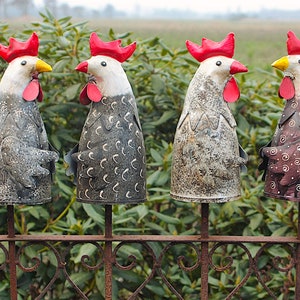 Könnte beinhalten: Vier Gartenornamente in Form von Metallhähnen. Jeder Hahn hat einen roten Kamm und Kehllappen, einen weißen Kopf und einen Körper mit einem einzigartigen Muster. Sie sind auf einem dekorativen Metallzaun mit grünem Hintergrund montiert.