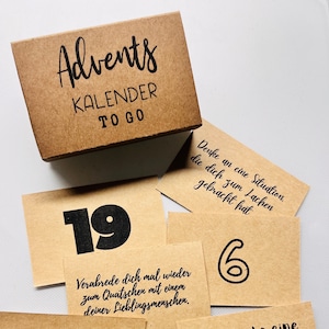 Könnte beinhalten: Eine braune Adventskalenderbox aus Pappe mit den Worten "Advents KALENDER TO GO" in Schwarz. Mehrere braune Karten mit schwarzen Zahlen und handschriftlichen Sätzen liegen um die Box herum.