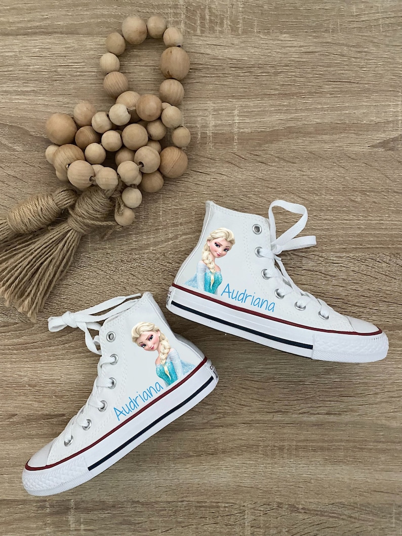 Puede incluir: Zapatillas blancas de lona de ca&ntilde;a alta con una imagen azul y blanca de Elsa de Frozen. Las zapatillas est&aacute;n personalizadas con el nombre "Audriana" en letras azules.